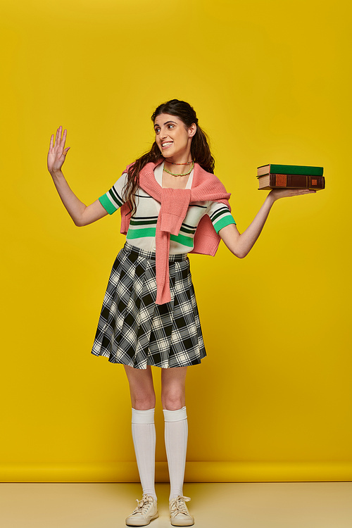 happy student with books, brunette woman in checkered skirt waving hand on yellow backdrop, brunette