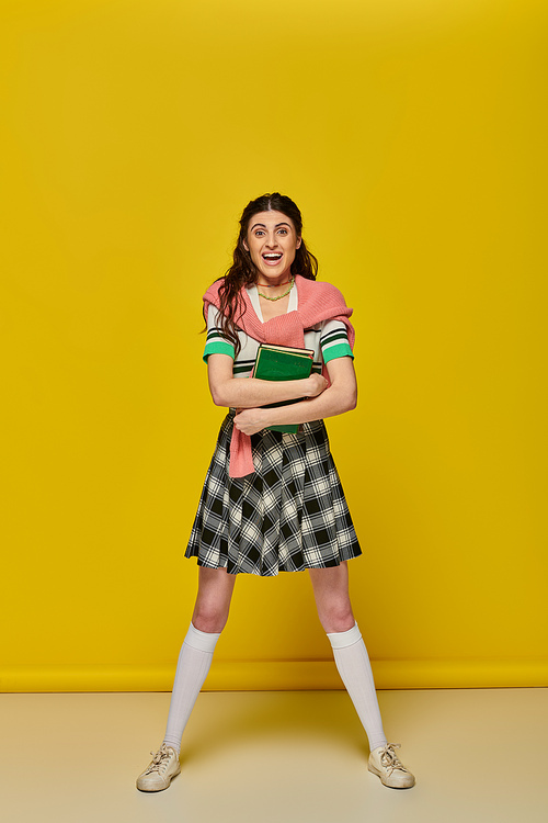 excited young woman in skirt standing with books on yellow backdrop, happy student, college outfit