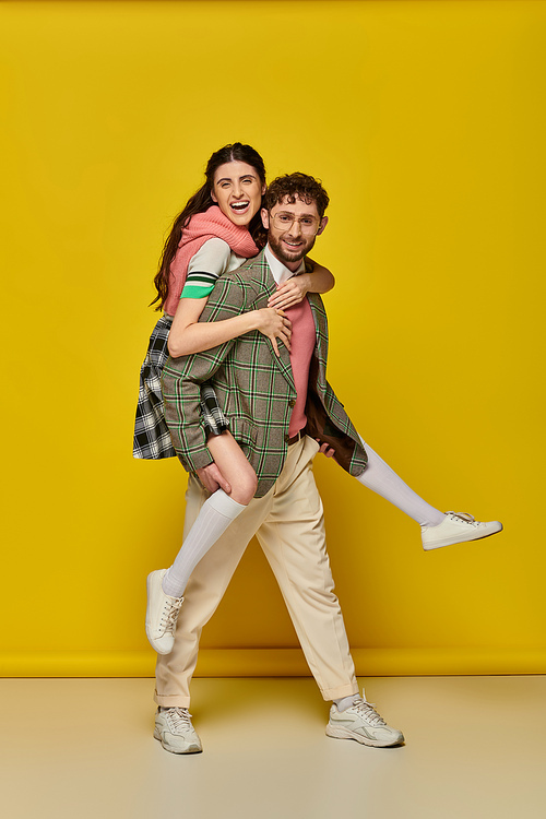 excited couple, bearded young man piggybacking brunette woman on yellow backdrop, funny students