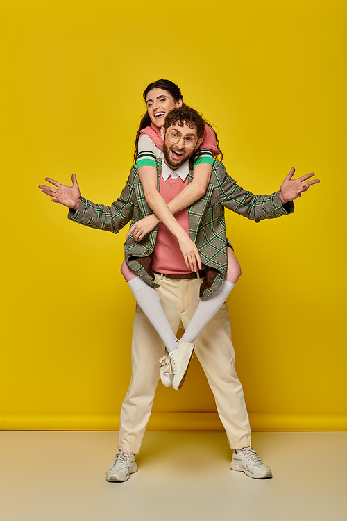 excited couple, funny, young man piggybacking brunette woman on yellow backdrop, student outfits