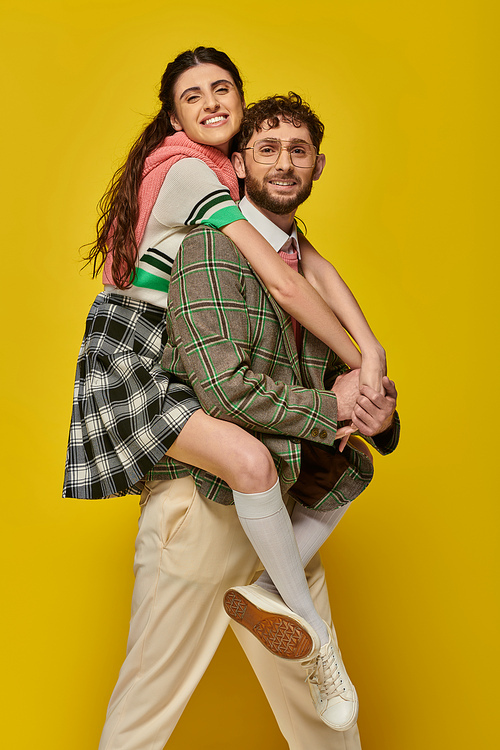 couple, funny students, happy man piggybacking young woman on yellow backdrop, college outfits