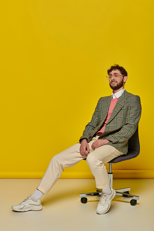 happy bearded man sitting on office chair, yellow backdrop, student in college outfit, glasses