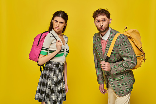 curious students standing with backpacks, looking at camera, yellow backdrop, college outfits, study