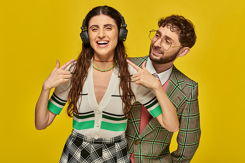 cheerful woman pointing at wireless headphones, listening music near bearded man, college students