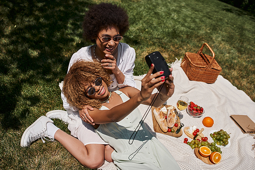 top view of joyful african american girlfriends taking selfie near food and wine, picnic in park