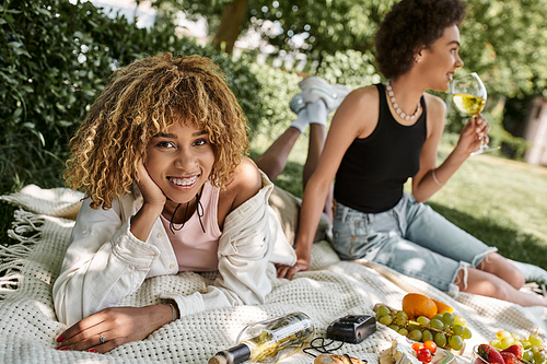 summer picnic, joyful african american woman looking at camera near girlfriend, wine and fruits