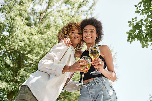 carefree african american girlfriends with wine glasses embracing in park, summer picnic