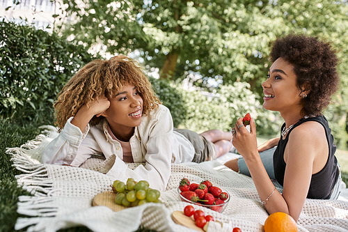 happy african American girlfriends talking near fresh fruits on blanket, summer picnic in park