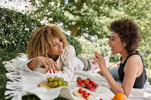 summer picnic, excited african american woman talking to girlfriend near fresh fruits on blanket