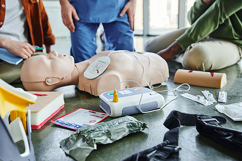 CPR manikin, defibrillator, wound care simulators, compression tourniquet, bandages and syringes near instructor and multiethnic participants of first aid seminar, emergency preparedness concept