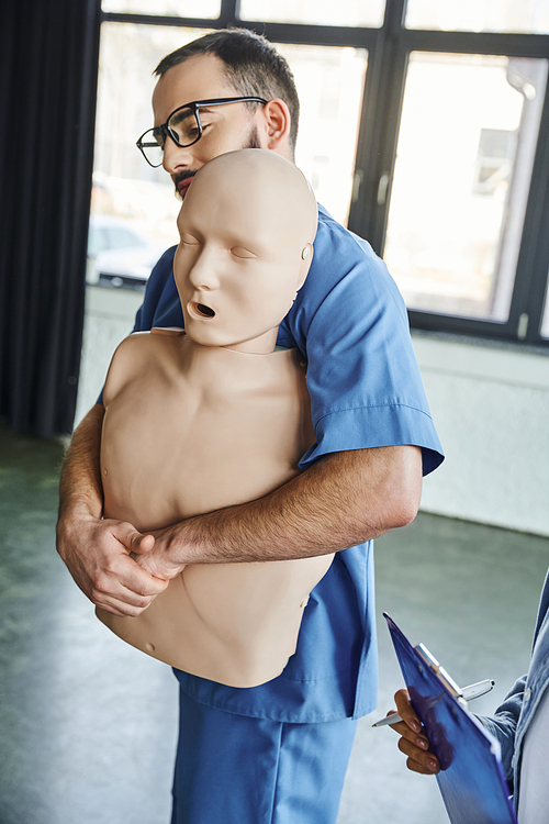 first aid seminar, young professional paramedic showing rescue techniques in case of chocking on CPR manikin near woman with clipboard and pen in training room, emergency situations preparedness
