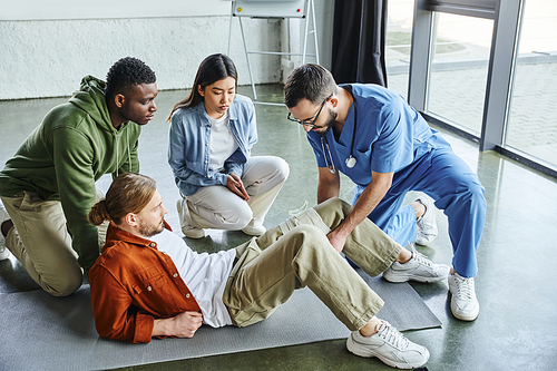african american man and asian woman looking at medical instructor applying compressive bandage on leg of participant of first aid training seminar, bleeding prevention techniques concept