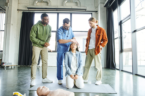 multiethnic man looking at professional paramedic bandaging head of asian woman near CPR manikin during first aid training seminar, importance of emergency preparedness concept