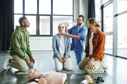 professional paramedic in eyeglasses and uniform bandaging head of asian woman near interracial men during medical seminal with CPR manikin and medical equipment, effective first aid concept