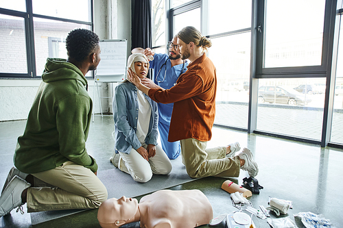 african american participant of first aid hands-on learning looking at young man and medical instructor bandaging head of asian woman near medical equipment, emergency preparedness concept