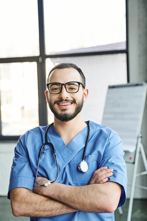 optimistic bearded paramedic in blue uniform and eyeglasses looking at camera and standing with folded arms and stethoscope near flip chart on blurred background, first aid training seminar concept