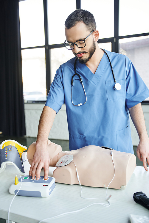 healthcare worker in blue uniform, stethoscope and eyeglasses operating automated defibrillator near CPR manikin, first aid hands-on learning and critical skills development concept