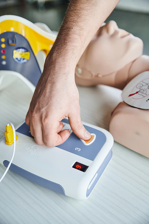 partial view of healthcare worker operating automated defibrillator while practicing cardiac resuscitation on CPR manikin, first aid hands-on learning and critical skills development concept