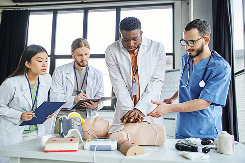 african american man in white coat practicing chest compressions on CPR manikin near paramedic, medical equipment and multiethnic students in training room, emergency situations response concept