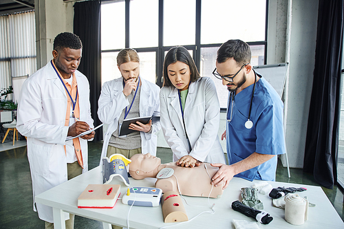 young asian student in white coat practicing chest compressions on CPR manikin near defibrillator, healthcare worker, and multicultural men with notebooks, emergency situations response concept