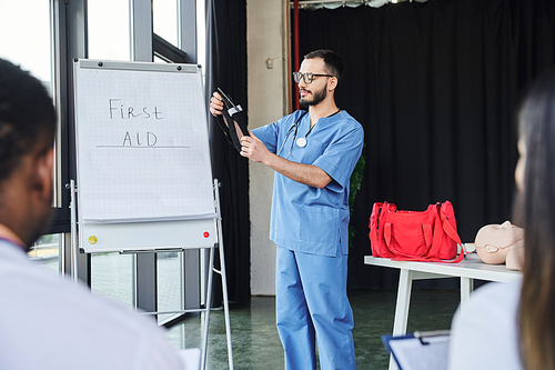 doctor in blue uniform and eyeglasses standing with compressive tourniquet near flip chart with first aid lettering and multiethnic students on blurred foreground, life-saving skills concept