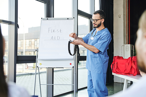 paramedic in eyeglasses and blue uniform standing at flip chart with first aid lettering and showing compressive tourniquet to students on blurred foreground, life-saving skills concept