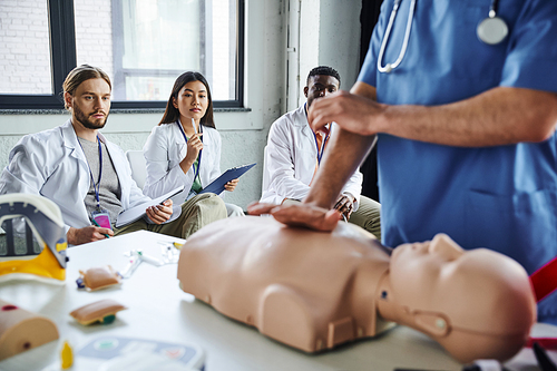 young multiethnic students in white coats looking at paramedic showing life-saving techniques on CPR manikin on blurred foreground, acquiring and practicing life-saving skills concept