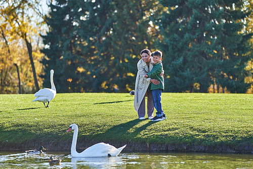 happy african american mother and son looking at swans in lake, nature, autumnal fashion, outerwear