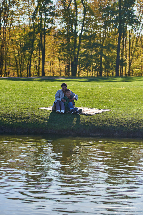 cheerful mother and son hugging in autumnal park, sitting on blanket near lake, african american