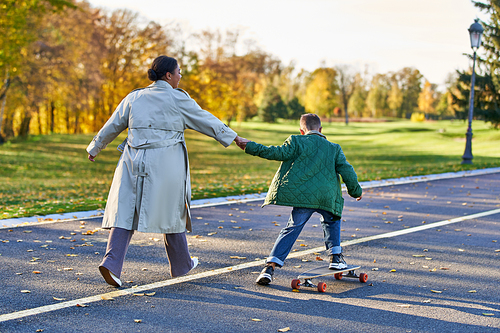 boy in outerwear riding penny board and holding hands with mother, african american,  autumn leaves