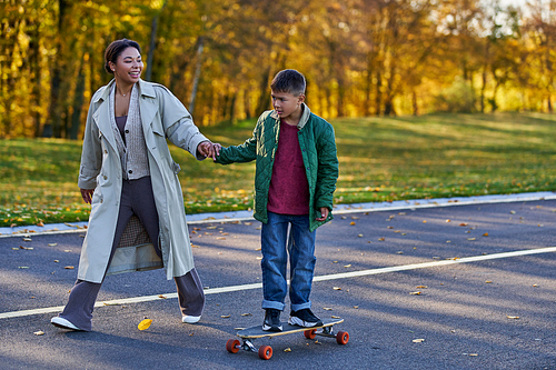 kid in outerwear riding penny board and holding hands with happy african american mom,  autumn