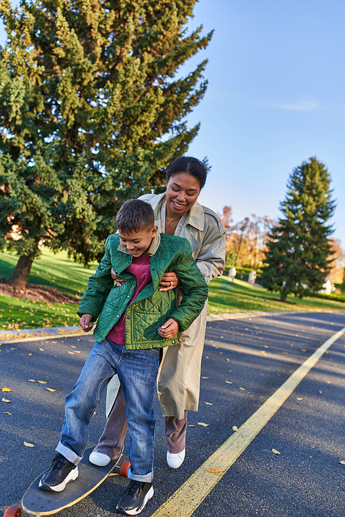 mother and son in autumn park, african american woman supporting boy on penny board, happy moments