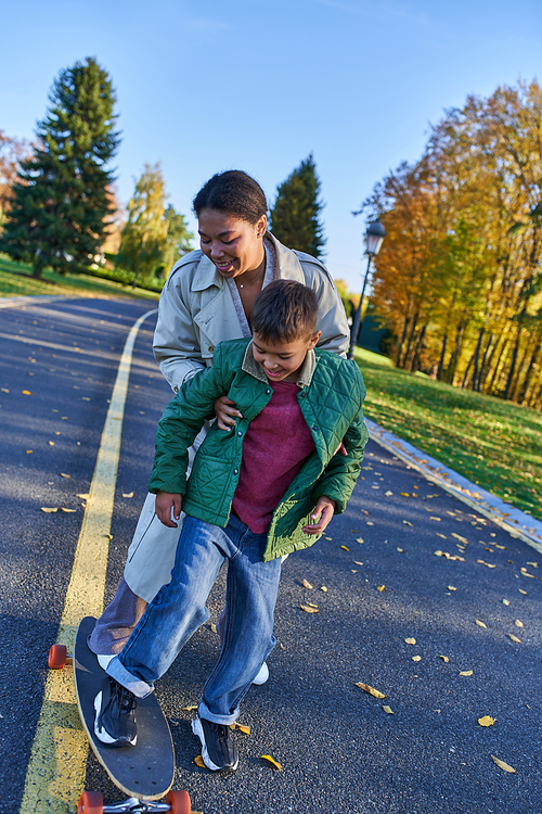 mother and son in autumn park, african american woman supporting boy on penny board, joyful moments