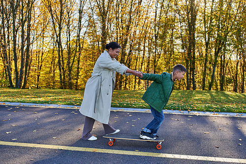 mother and son in autumn park, happy african american woman holding hands with boy on penny board