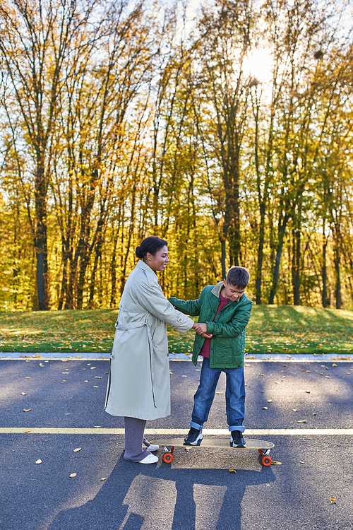 mother and son in park, happy african american woman holding hands with boy on penny board, autumn