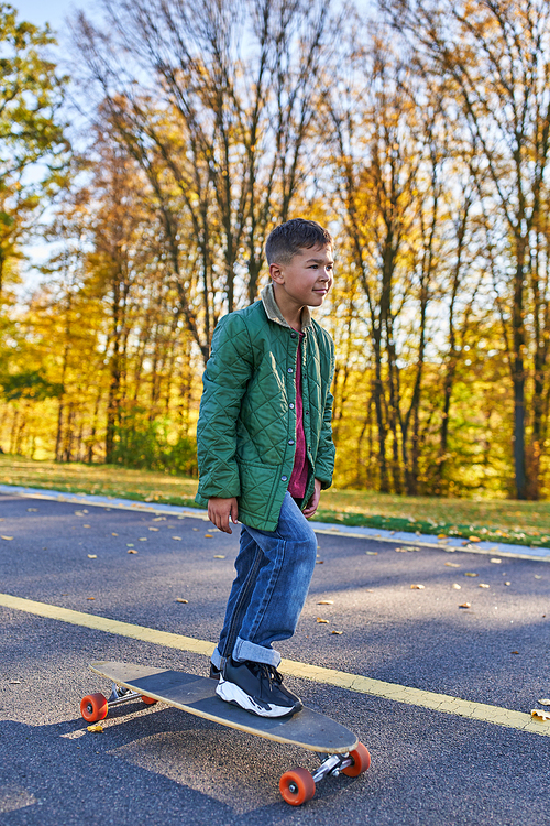 preteen african american boy in outerwear and jeans riding penny board, autumn park, fall season