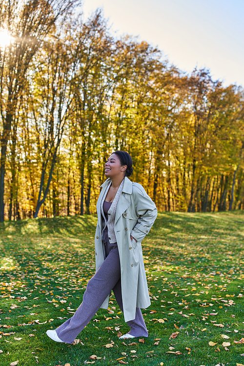happy african american woman in trench coat walking on grass with fallen leaves, autumn, fashion