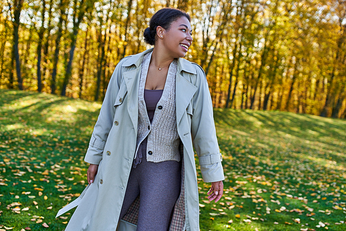 cheerful african american woman in trench coat walking in autumn park, fashion outfit, style