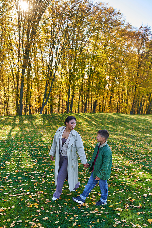 cheerful african american woman holding hands with son, walking on grass with golden leaves, autumn