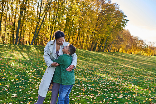 happy african american woman hugging with son, standing on grass with golden leaves, autumn