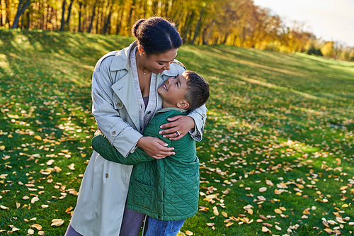 happy african american woman hugging with son, standing near golden leaves, autumn, motherly love