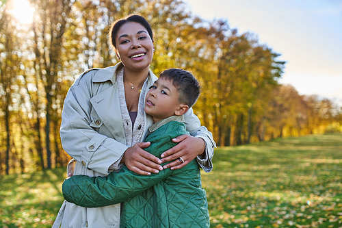 motherly love, joy, african american woman hugging cute son, standing near golden leaves, autumn