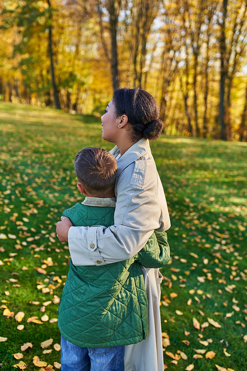 motherly love, back view, african american woman hugging cute son, standing near golden leaves