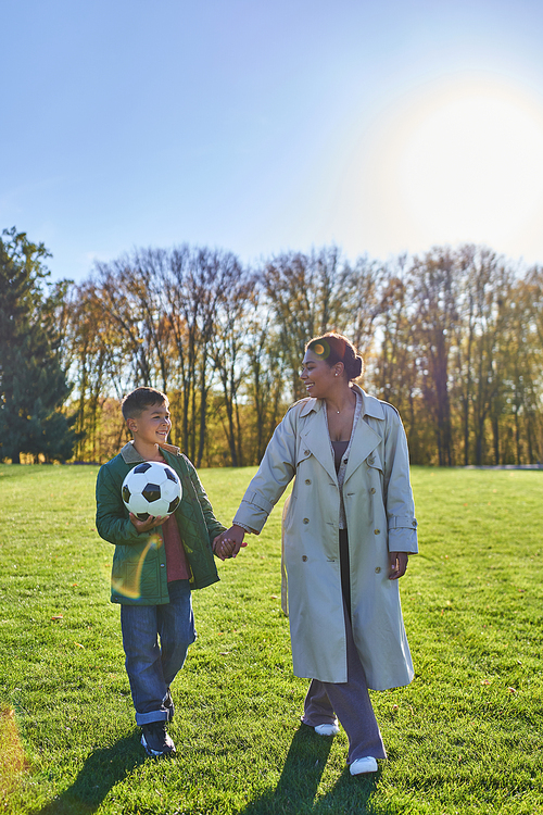 african american boy holding soccer ball, walking with mom on green field, holding hands, autumn