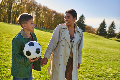 happy african american boy holding soccer ball, walking with mom on green field, outerwear, autumn