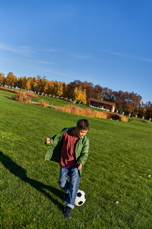 sporty african american boy in autumnal outerwear playing football on green field, soccer, autumn