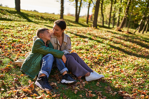 motherly love, cheerful african american woman and son sitting on grass with golden leaves, autumn