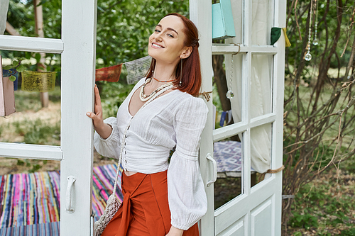 positive and trendy redhead woman in boho styled outfit standing outdoors in retreat center