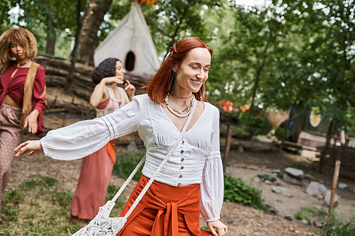 cheerful redhead woman in boho outfit spending time near multiethnic women in retreat center