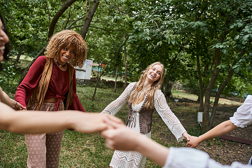 happy multiethnic girlfriends in boho clothes holding hands and dancing in retreat center